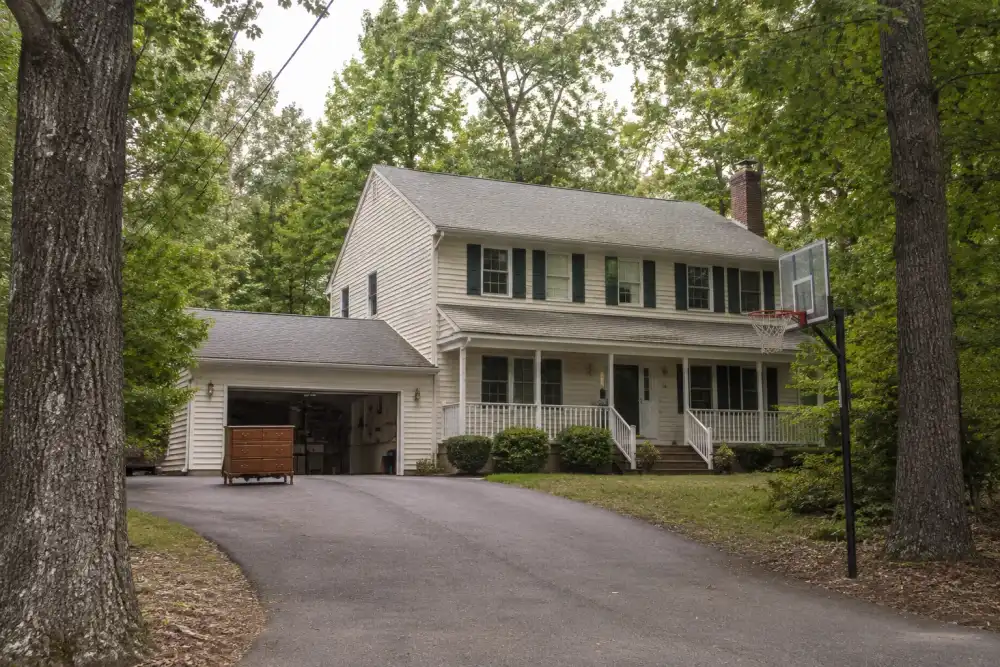 Illustrative scene: chesterfield-inspired two-story colonial home with a sloping driveway, mature oak trees and a basketball hoop, old dresser staged near open garage