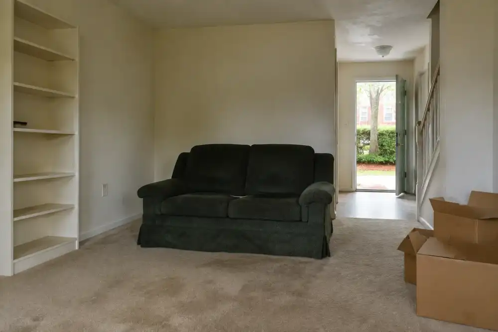 Illustrative scene: vacant modest townhouse interior with a worn loveseat, empty shelving and abandoned cardboard boxes, daylight from an open front door, no papers or people
