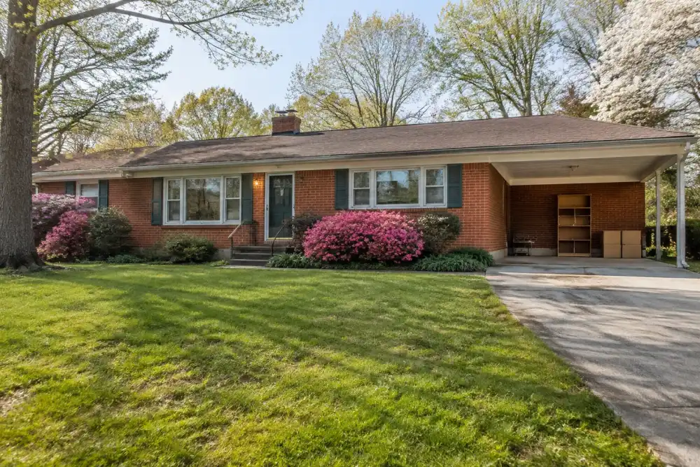 Illustrative scene: henrico-inspired mid-century single-story brick ranch with a broad front lawn, carport and azaleas, small shelving unit and boxes ready for pickup in carport