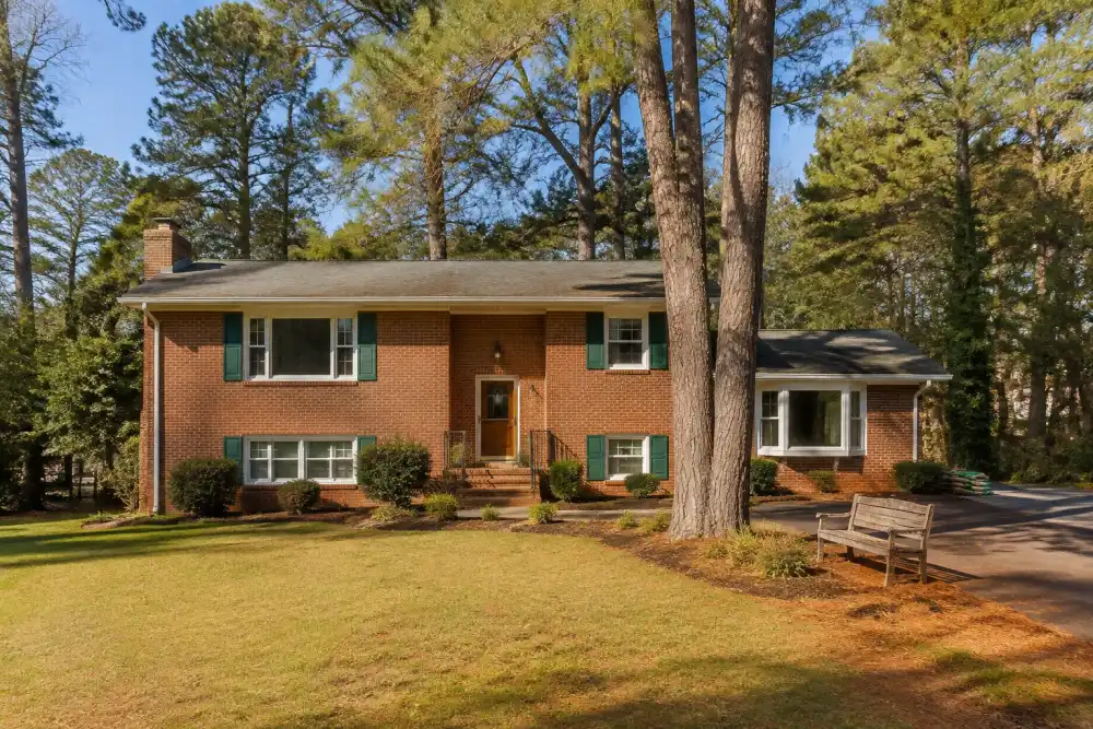 Illustrative scene: mechanicsville-inspired modest red-brick split-level house with a side driveway and tall pine trees, old garden bench and bags beside driveway