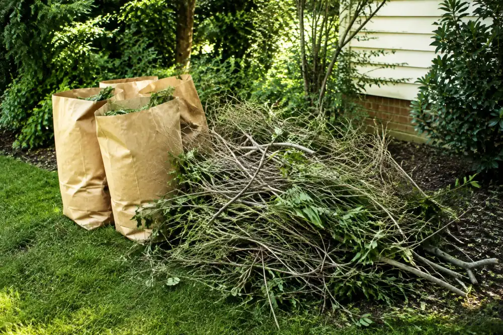 Illustrative scene: pile of trimmed branches, bundled twigs and brown paper yard bags beside a suburban garden, dappled green shade, no power tools